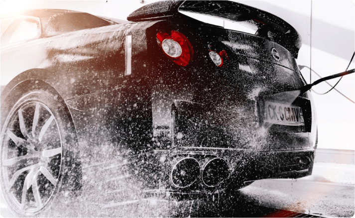A black sports car being washed with high-pressure water, capturing the cleaning process with water droplets and sunlight reflections.